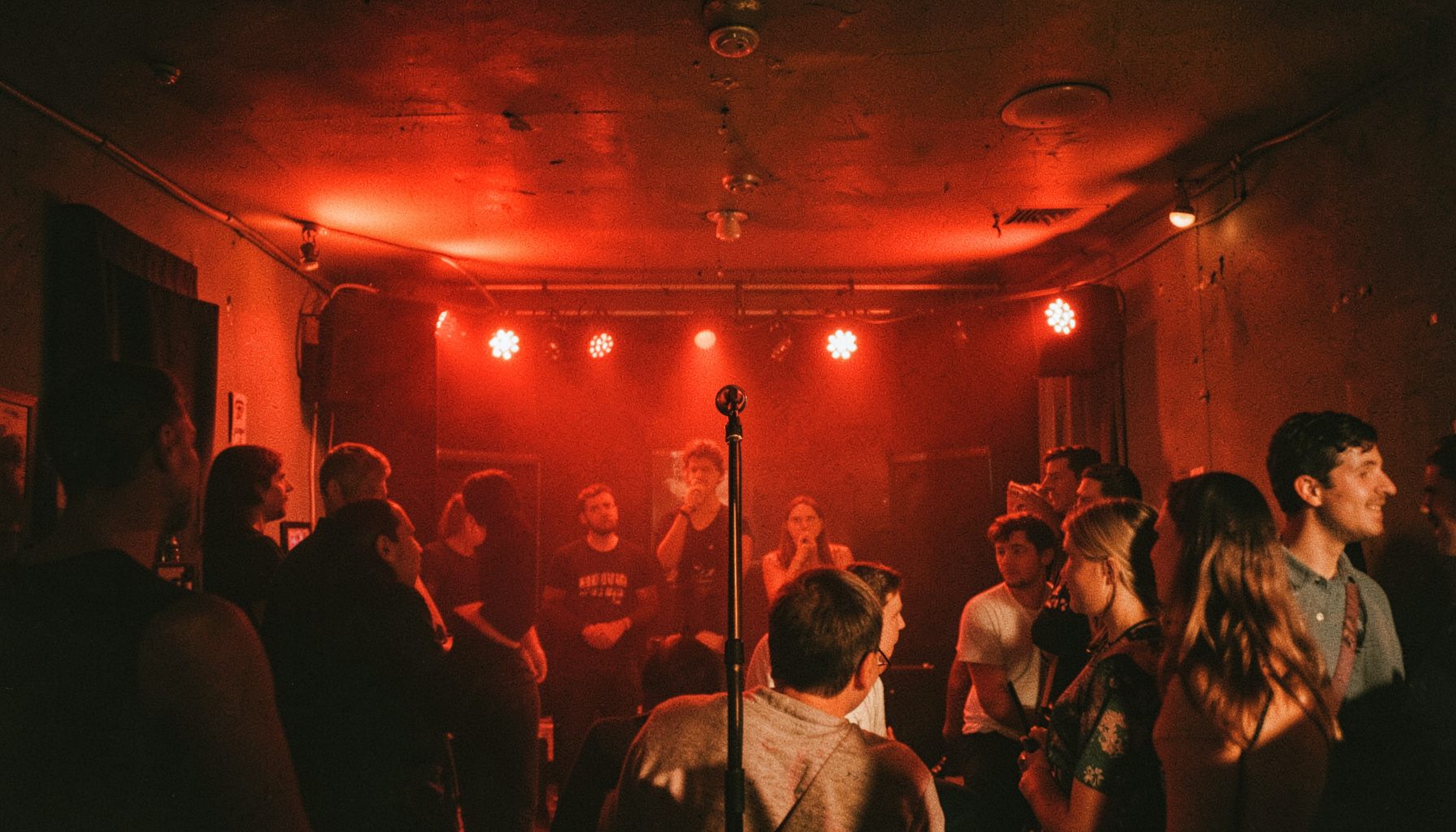 Interior of a small club with red stage lights reflecting off a low ceiling, microphone stand center stage