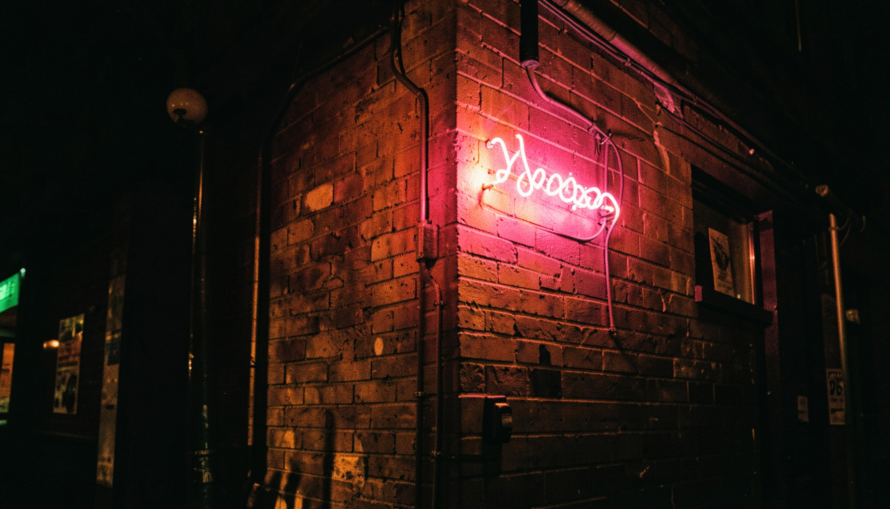 Neon sign glowing pink against a dark brick wall outside a small music venue at night