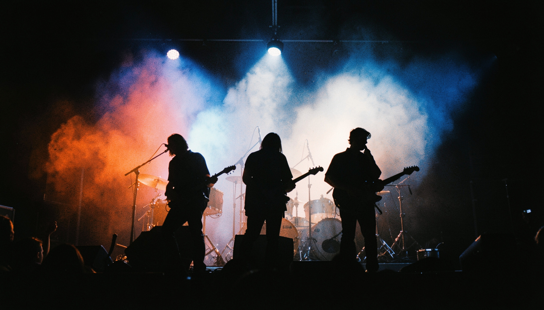 Silhouetted band members on a dark stage under deep blue and white spotlights, haze in the air