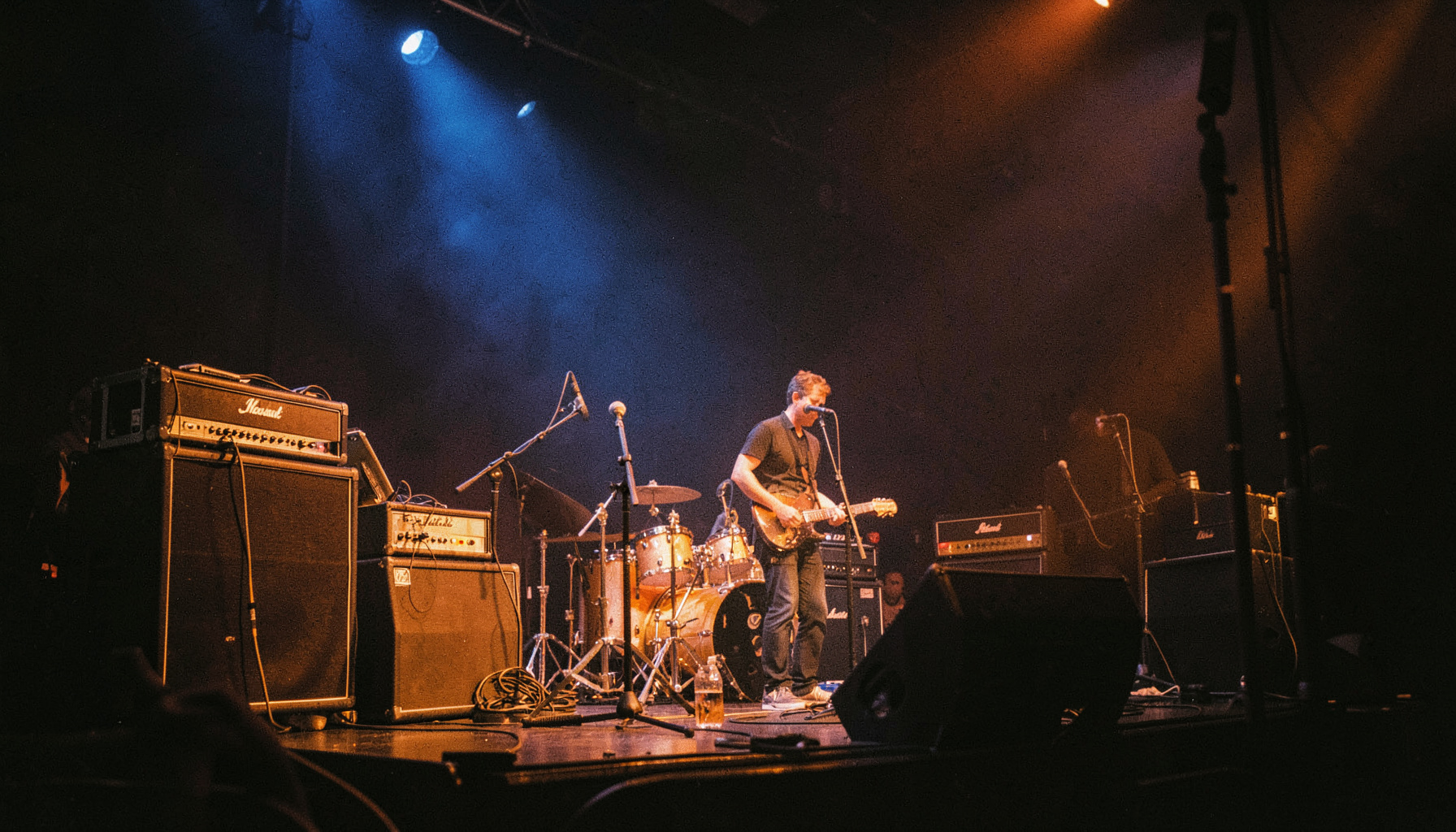 Dimly lit stage with amplifiers and microphone stands, deep blue and amber lighting, moody concert atmosphere