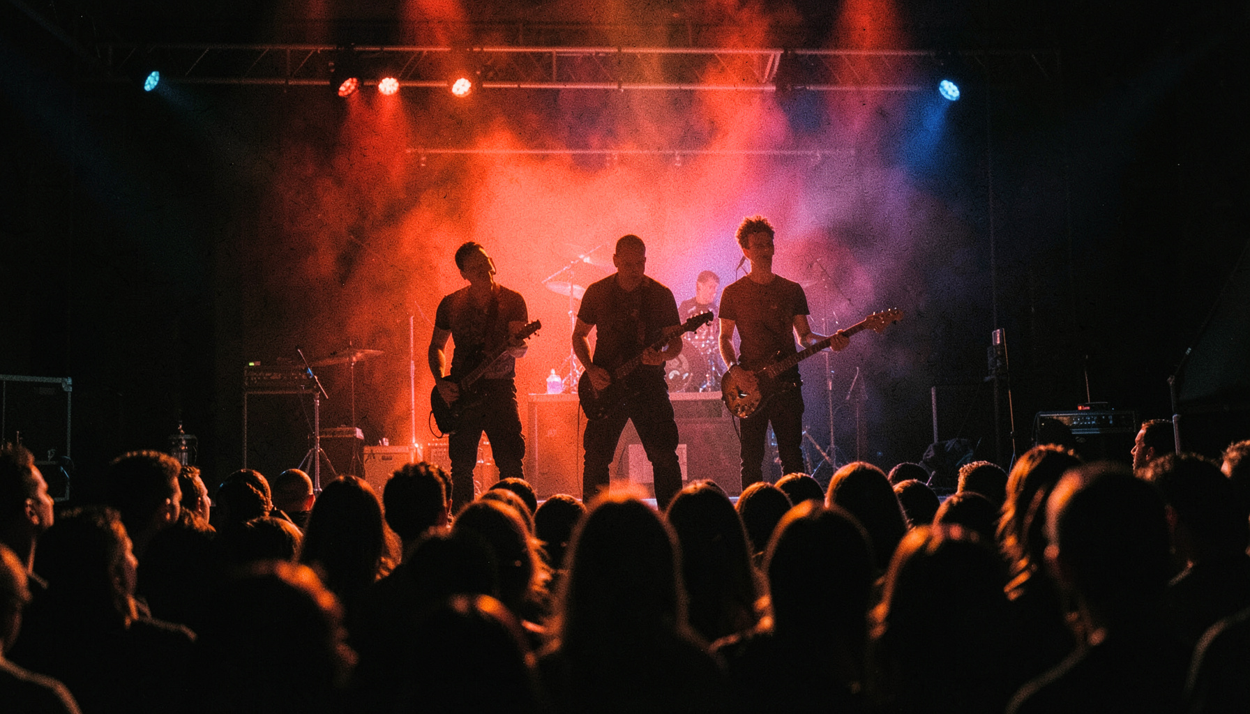 Silhouetted band members on a small stage bathed in deep red and blue light, crowd visible in the foreground