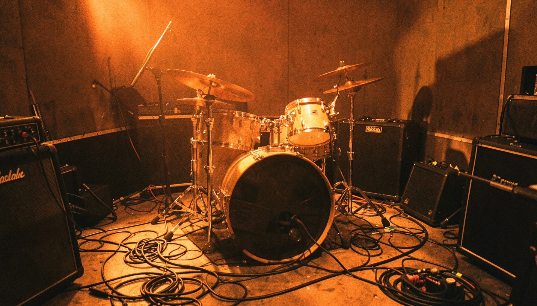 Guitar and drum kit in a dimly lit rehearsal space, warm tungsten glow, cables and amps scattered across the floor