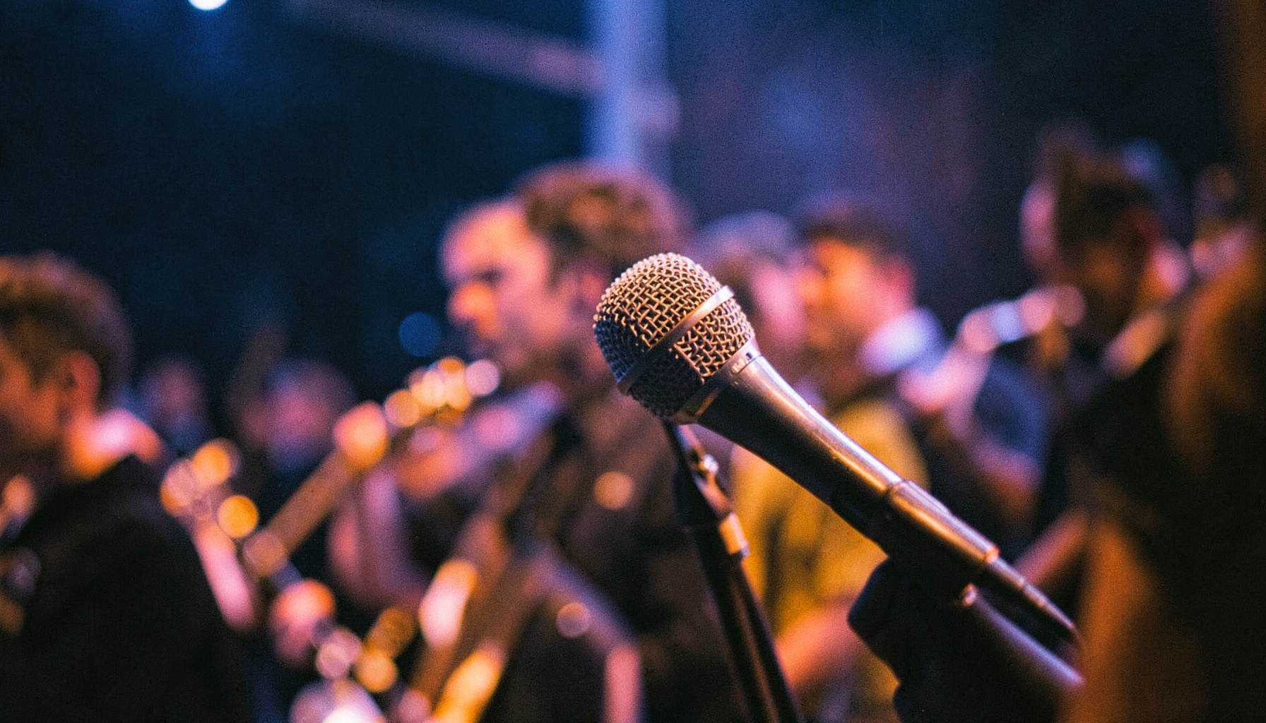 Close-up of a microphone on stage with blurred band members in the background, moody blue and purple lighting
