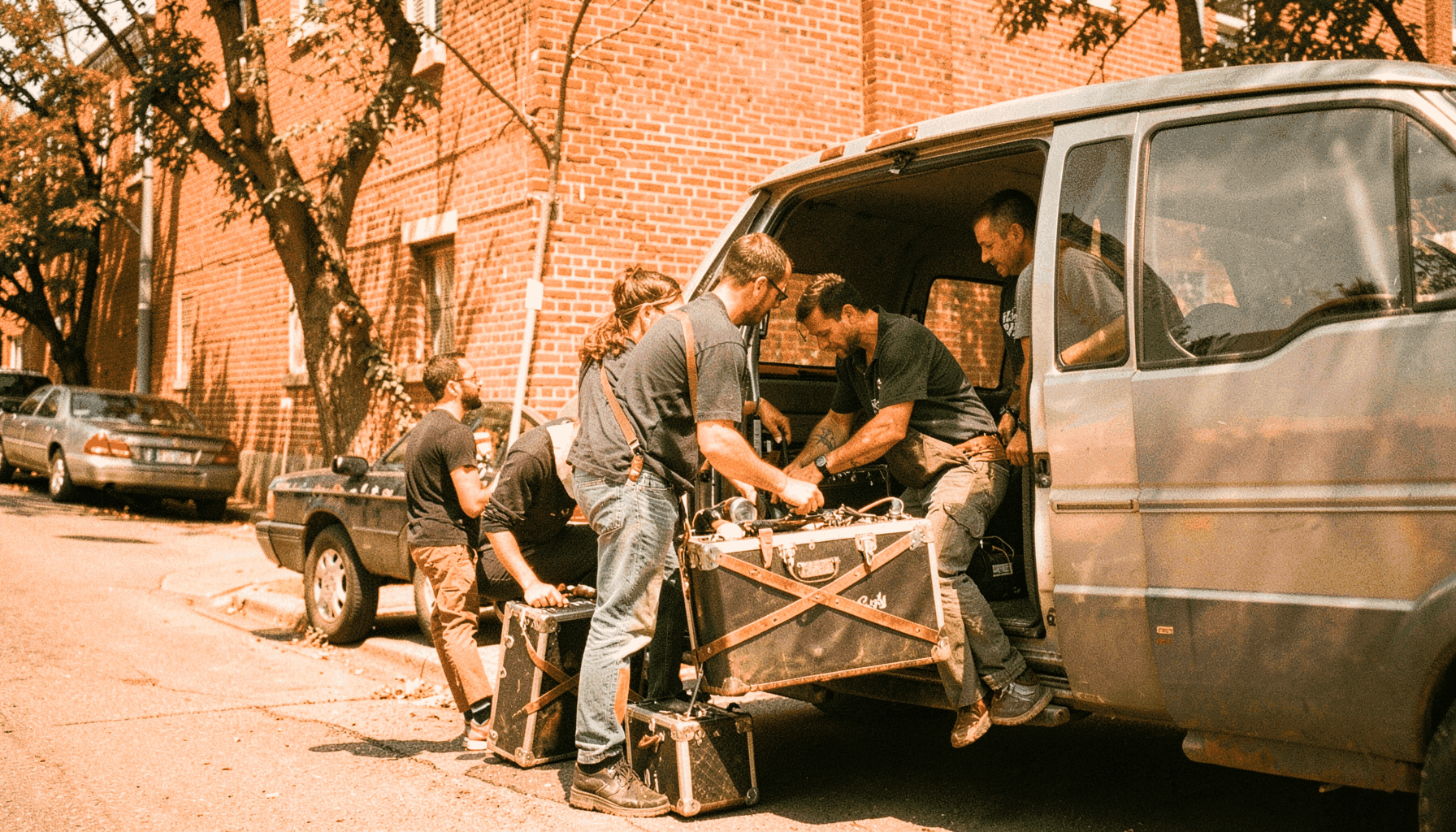 Band members loading gear into a van on a sunny afternoon, brick wall and street in the background