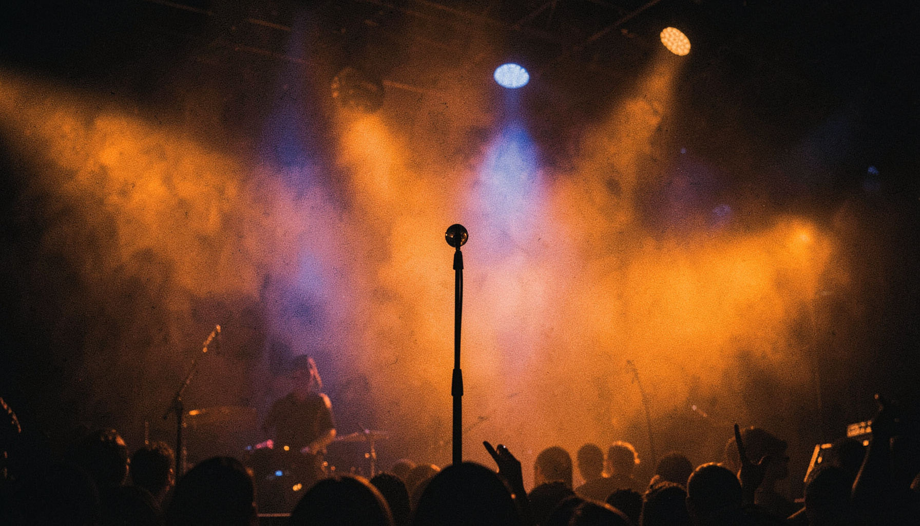 Dimly lit stage with warm amber and deep blue stage lights cutting through haze, microphone stand in silhouette, concert atmosphere